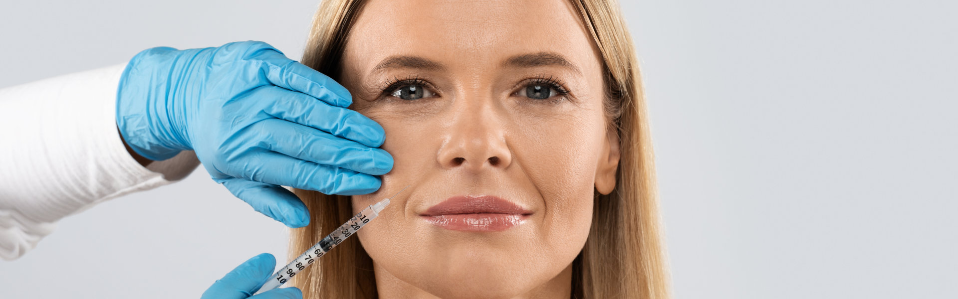 A woman receiving a cosmetic injection in her cheek, administered by a professional wearing blue gloves. She has a neutral expression.